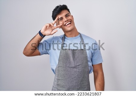 Hispanic young man wearing apron over white background doing peace symbol with fingers over face, smiling cheerful showing victory 