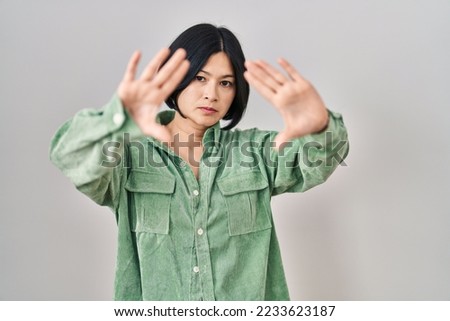 Young asian woman standing over white background doing frame using hands palms and fingers, camera perspective 