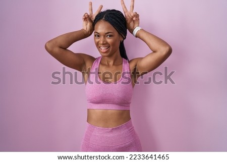 African american woman with braids wearing sportswear over pink background posing funny with fingers on head as bunny ears, smiling cheerful 