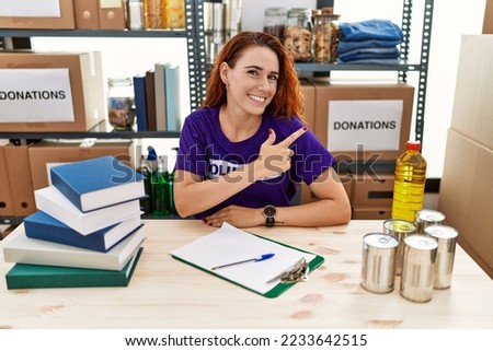 Young redhead woman wearing volunteer t shirt at donations stand cheerful with a smile on face pointing with hand and finger up to the side with happy and natural expression 