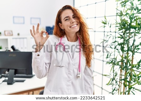 Young redhead woman wearing doctor uniform and stethoscope at the clinic smiling positive doing ok sign with hand and fingers. successful expression. 