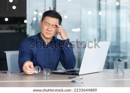 Asian businessman boss sick has severe headache, man in glasses sitting at desk using laptop at work inside office, migraine office worker investor overtired. Royalty-Free Stock Photo #2233644889