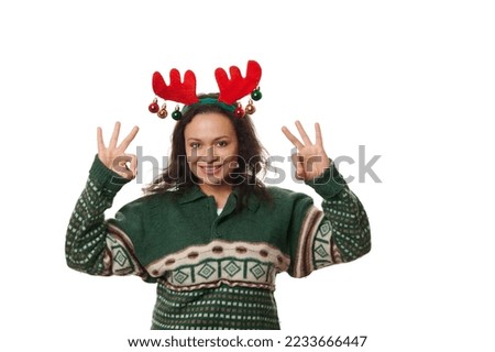 Latin American beautiful woman, wearing a warm woolen green Xmas patterned sweater and deer antler hoop, gesturing, showing OK sign, cutely smiling looking at camera on white background. Copy ad space