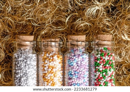 Nonpareils in small glass bottles, over golden angel hair. Winter and christmas colored mixes of edible confectionery of tiny sugar balls. Hundreds and Thousands, used as decoration and as topping.