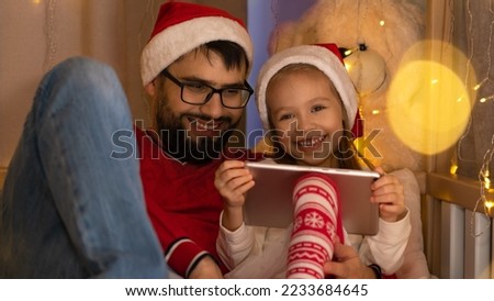 Father and little girl in red santa hats use digital tablet and laugh. Happy family spend New Years Christmas holiday time.