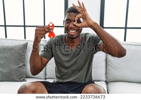 Young african american man holding support red ribbon sitting on the sofa smiling happy doing ok sign with hand on eye looking through fingers 