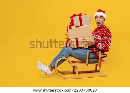 Full body side view fun merry young man wear red knitted Christmas sweater Santa hat posing sledding hold many present boxes isolated on plain yellow background. Happy New Year 2023 holiday concept