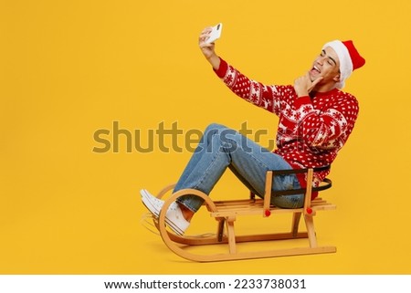 Full body merry young man wear red knitted Christmas sweater Santa hat posing sledding doing selfie shot on mobile cell phone isolated on plain yellow background. Happy New Year 2023 holiday concept