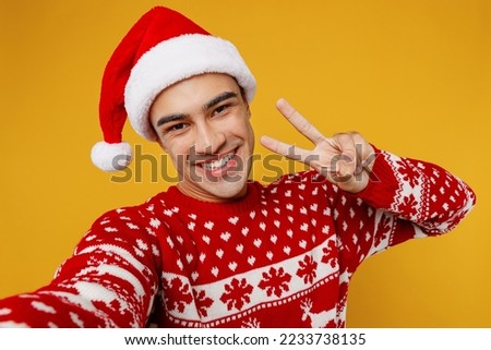 Close up merry young man wear red knitted christmas sweater Santa hat posing do selfie shot pov on mobile cell phone show v-sign isolated on plain yellow background Happy New Year 2023 holiday concept
