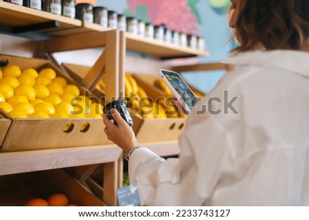 Close-up side view of unrecognizable young woman choosing products in market walking along counters taking photos of items using smartphone. Female looking up nutrition facts using mobile phone.