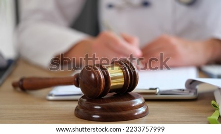 Wooden gavel of judge on table close-up, and blurred doctor doing paperwork in medical clinic. Litigation, malpractice and forensic concept. Royalty-Free Stock Photo #2233757999