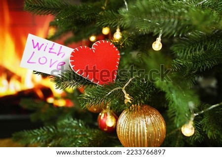 red heart and a sign with the inscription With Love on a Christmas tree against the background of a fireplace. Valentine's Day