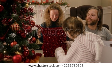 Happy caucasian family opening gifts on Christmas under decorated Christmas tree. Mother unpacks red festive sparkly dress. Warm atmosphere at home on Christmas or New Year. Winter holidays.