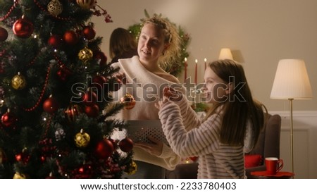 Happy young girl with mom decorating Christmas tree with balls and toys. Dad hangs up Christmas wreath on the wall. Family preparing home for winter holidays or New Year. Slow motion.