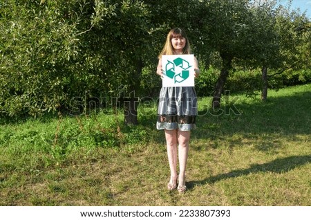 Smiling volunteer young woman in grey dress holding recycling sign on natural background for green future for planet.