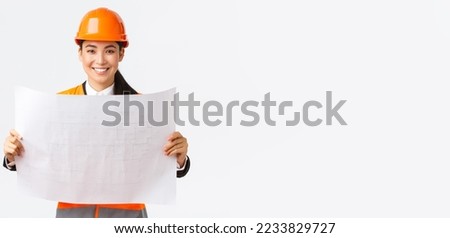 Excited smiling asian female engineer at construction area study blueprints, looking pleased while reading documents, building plan, standing in safety helmet over white background.
