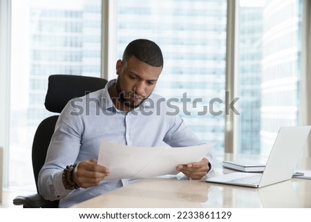 Focused busy African business leader man reading paper document, sales report, boss order, doing paperwork at workplace. Legal expert, CEO, manager checking contract at laptop Royalty-Free Stock Photo #2233861129