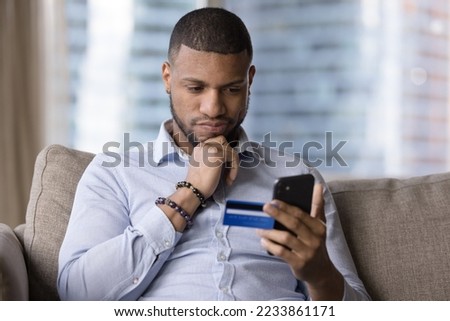 Thoughtful African credit card user holding mobile phone, looking at screen, touching chin in doubts, making decision, using ecommerce banking app for making payment, shopping Royalty-Free Stock Photo #2233861171