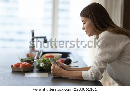 Serious focused chef girl using mobile phone at kitchen table, watching cook blogger video recipe, standing at table with fresh vegetables, natural food ingredients for salad, dinner
