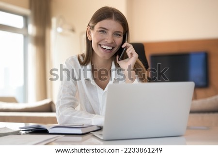 Happy young business woman working from home, speaking on cellphone at laptop, sitting at table, making mobile phone call, talking, listening, laughing, looking at camera. Female head shot portrait