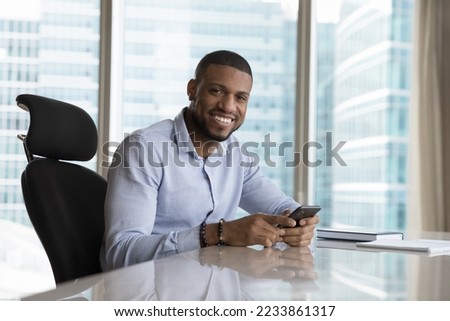 Happy confident African American business owner man holding mobile phone at office workplace, looking at camera with toothy smile. Corporate portrait of smartphone user at work desk