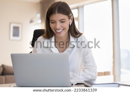 Happy successful freelance employee girl with wireless earphone in ear using laptop, smiling, talking to client on video call at work table. College student watching learning webinar on Internet