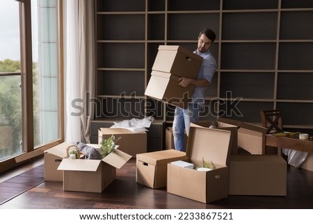 Busy serious renter man carrying stack of paper cardboard relocation boxes in empty room with pile of containers, leaving apartment, moving into new flat. Wide shot, full length Royalty-Free Stock Photo #2233867521