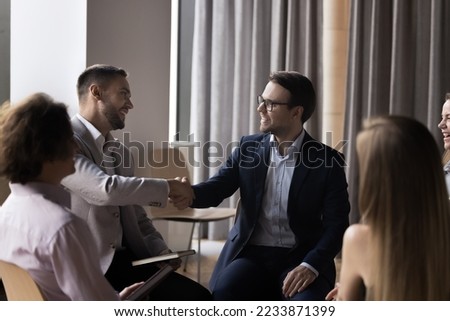 Two businessmen in formal suits shaking hands, start group formal meeting in office, gathered to discuss business, making profitable commercial deal. Express respect, negotiation process, etiquette