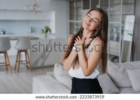 Attractive Swedish young adult woman in white blouse and black pants folded hands on chest eyes closed feel gratitude enjoys weekend at new home. Stunning blonde girl feels goos after massage.  Royalty-Free Stock Photo #2233873959