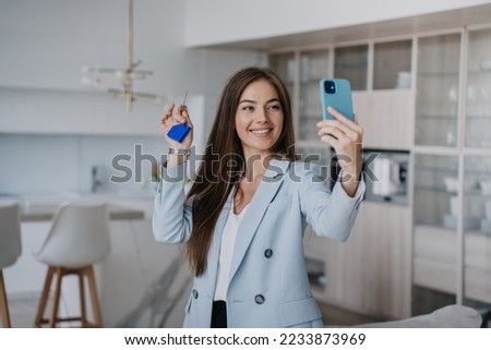 Pretty beautiful young woman in blue jacket shows keys of a new house to parents during video call by phone. Excited blonde businesswoman screams in happiness, purchased a flat. Realtor holds keys.