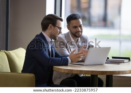 Two young businessmen in formal suits discuss new software sit at table with laptop in workspace, work on startup project, manager offering, presenting new services to client on device meet in office Royalty-Free Stock Photo #2233875957