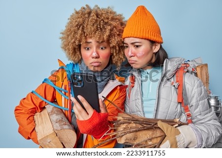 Displeased two women friends focused at smartphone screen check newsfeed and make selfie carry wood feel embarrassed dressed in comfortable clothes isolated over blue background. Hiking concept