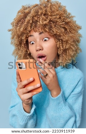 Photo of shocked curly haired woman stares impressed at smartphone screen reads news with stupefied expression wears casual jumper receieves message isolated over blue background. Omg thats terrible