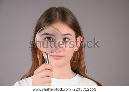 Teenage girl with magnifying glass on gray background. Female eyes with strabismus.  Royalty-Free Stock Photo #2233912651