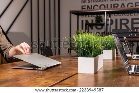 End of workday, finishing work in office concept. Hand closing laptop computer lid in modern interior with wood desk and plants. High quality photo