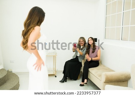 Cheerful mom taking a photo of her daughter shopping for a wedding gown at the bridal store