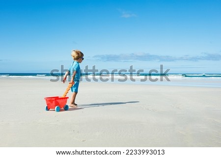 Happy little boy in sunglasses walk on the white sand beach playing with toy cart view from the back