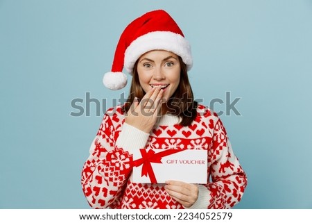 Young merry woman wear red sweater Santa hat posing hold store gift certificate coupon voucher card cover mouth isolated on plain pastel light blue cyan background. Happy New Year 2023 holiday concept