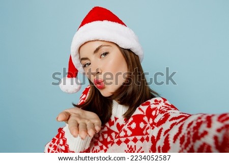 Close up young merry woman wear red sweater Santa hat posing do selfie shot pov mobile cell phone blow air kiss isolated on plain pastel light blue cyan background Happy New Year 2023 holiday concept