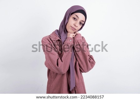 Charming serious young beautiful woman wearing hijab and pink overshirt over white background keeps hands near face smiles tenderly at camera