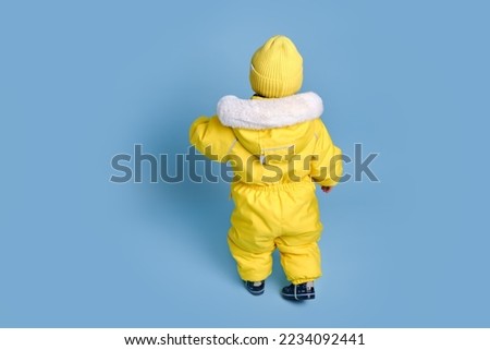 Happy toddler baby in winter clothes snowsuit on studio blue background. A child in a warm yellow jumpsuit with a hood. Kid aged one year five months Royalty-Free Stock Photo #2234092441
