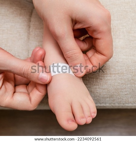 Mother woman sticks a medical adhesive plaster on the toddler baby leg. Mom s hand with sticky wound protection tape and child s foot. Kid aged one year and three months