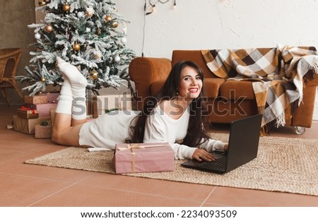 A happy girl in a Santa hat wishes her friends Merry Christmas in a video chat on a laptop. A young woman lying under a Christmas tree among gift boxes in a home interior