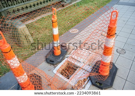 Open cover on a sidewalk access panel with hazard cones and fencing.