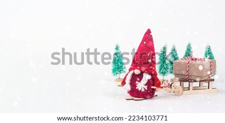 Scandinavian dwarf Nisse in red clothes, with skis in his hands, with pigtails sticking out. The gnome is carrying a sled with a gift.There are Christmas trees in the background.