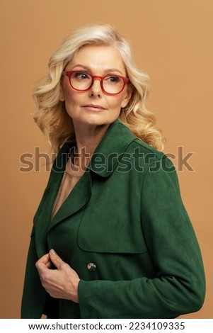 Confident mature fashion model wearing red eyeglasses, stylish green jacket posing for pictures isolated on background 