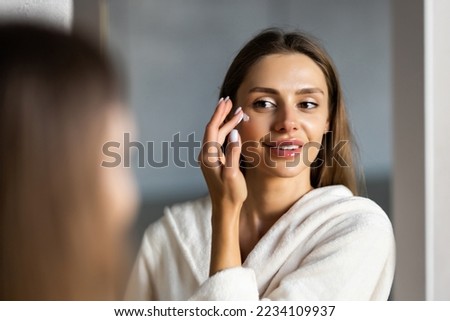 Close up cropped portrait of a young beautiful woman in bath towel smiling applying face body cream on her face for rejuvenation soft moisturizing effect. Royalty-Free Stock Photo #2234109937