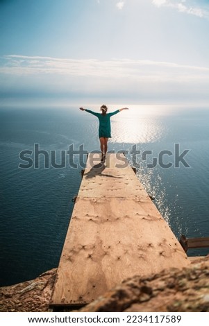 A girl sits on a wooden springboard for jumping with a rope. In a dark green dress with her hands in the air. Royalty-Free Stock Photo #2234117589