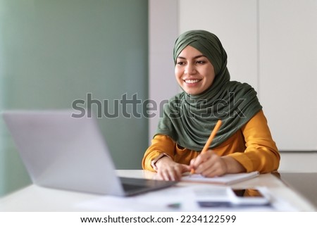Young Muslim Woman Watching Webinar On Laptop At Home And Taking Notes, Smiling Arab Lady In Hijab Study Online With Computer While Sitting At Desk Indoors, Enjoying Distance Learning, Free Space