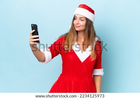 Young caucasian woman dressed as mama noel isolated on blue background making a selfie